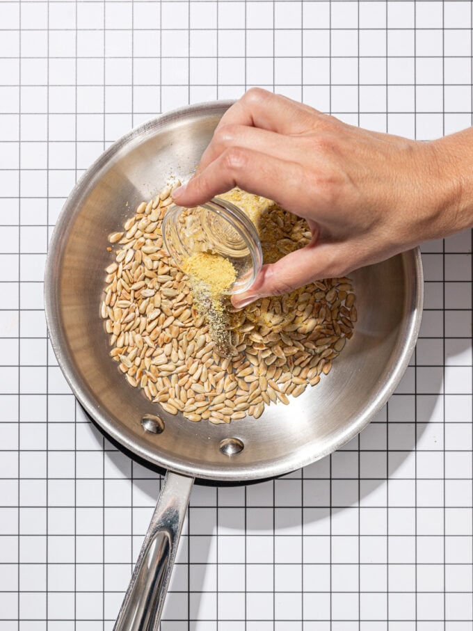 hand pouring garlic powder onto skillet of sunflower seeds