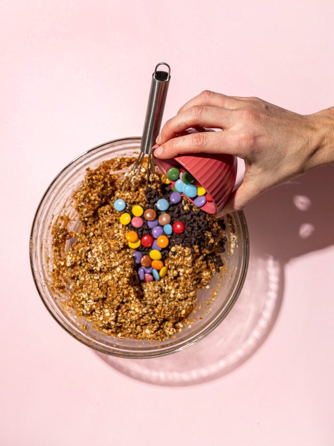 hand pouring colourful candies into monster cookie batter in glass bowl